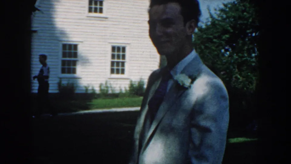 The suited man appears partly in shadow near a white clapboard building and lawn in the 1959 Nebraska home-movie scene.