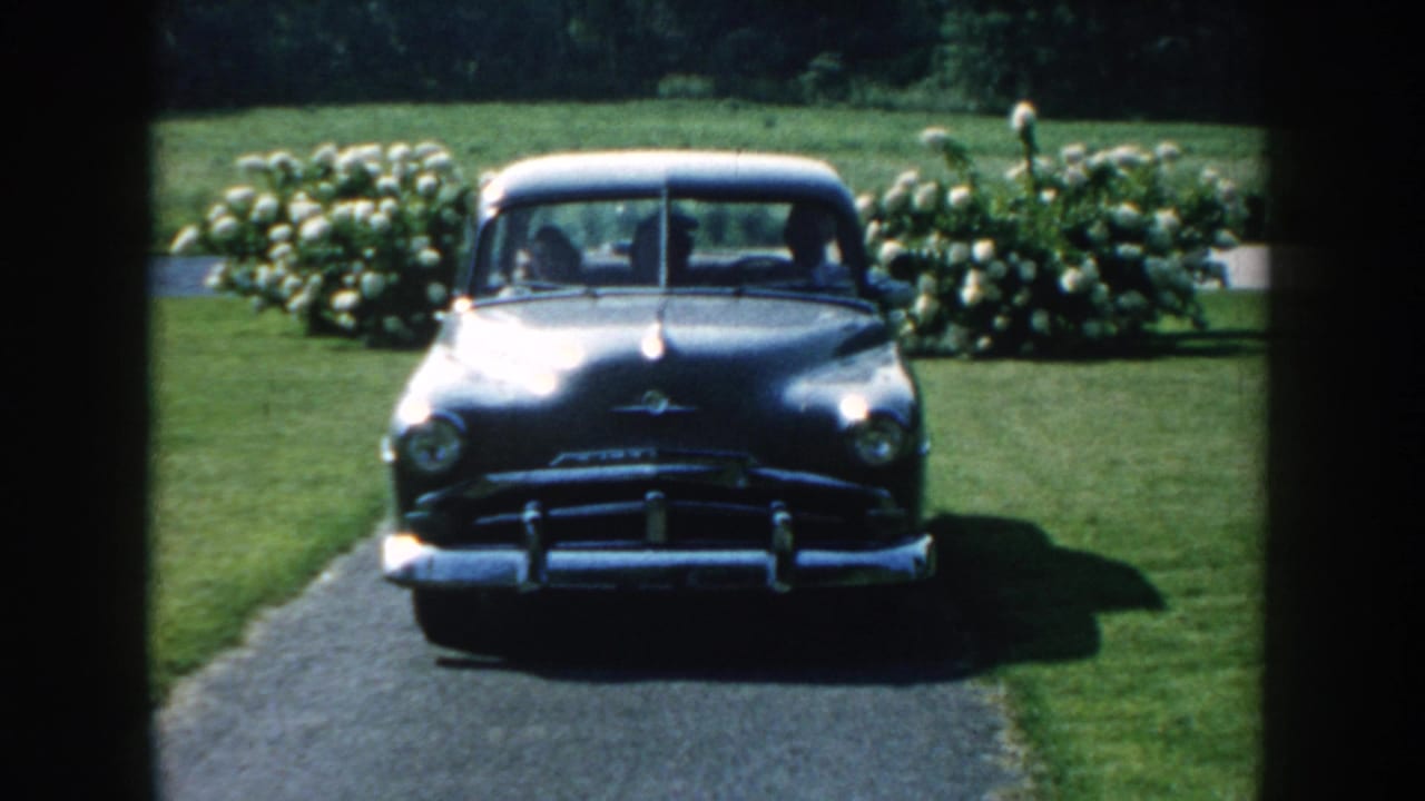 1959 Nebraska 8mm frame showing a dark classic sedan stopped or approaching on a residential driveway bordered by lawn and hydrangea-like bushes.