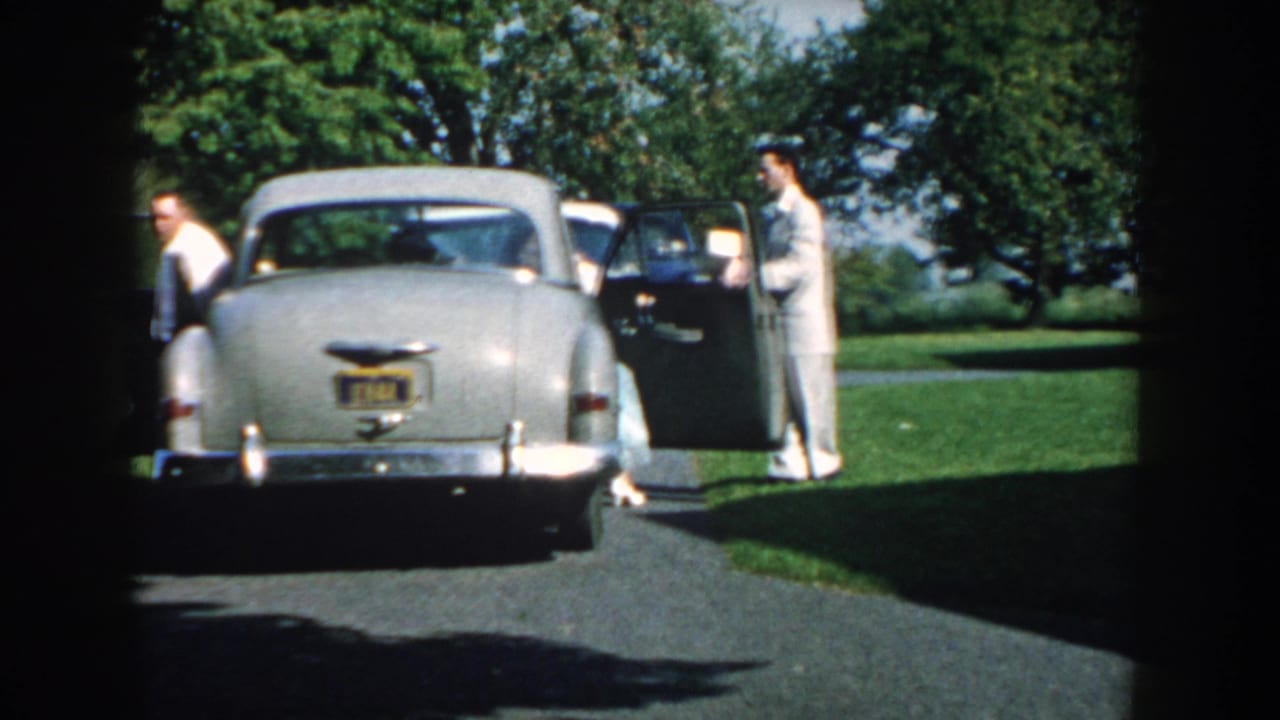 1959 Nebraska home-movie frame with two adult men by a light gray sedan, one door open, in a sunny residential outdoor setting.