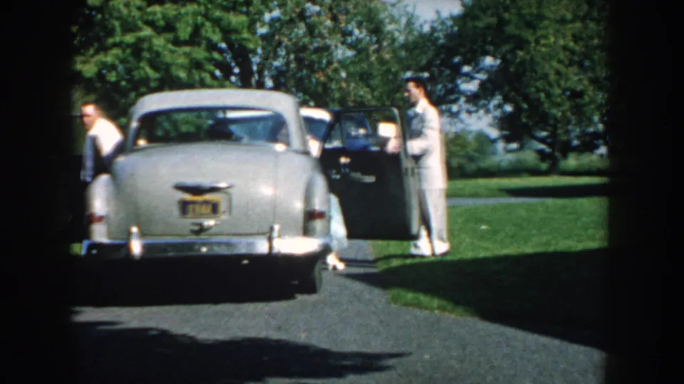 1959 Nebraska home-movie frame with two adult men by a light gray sedan, one door open, in a sunny residential outdoor setting.