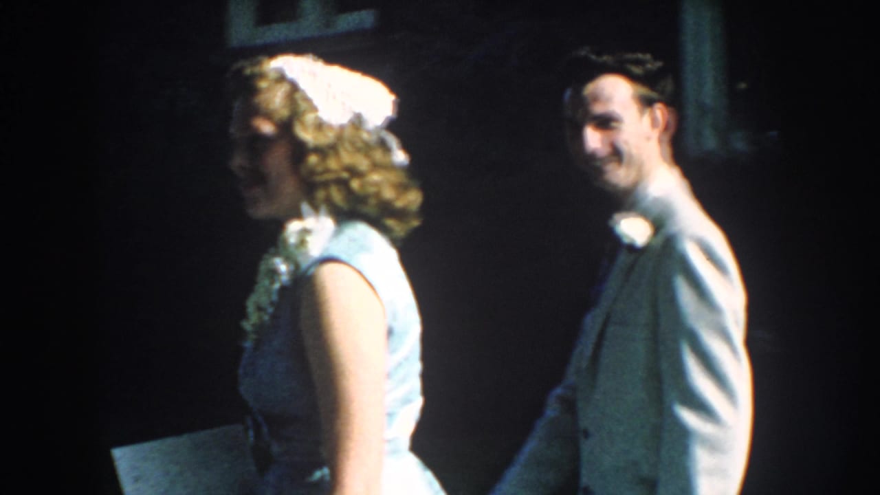 1959 Nebraska home-movie frame of a woman in a light blue dress and white headband near a smiling man in formal attire.