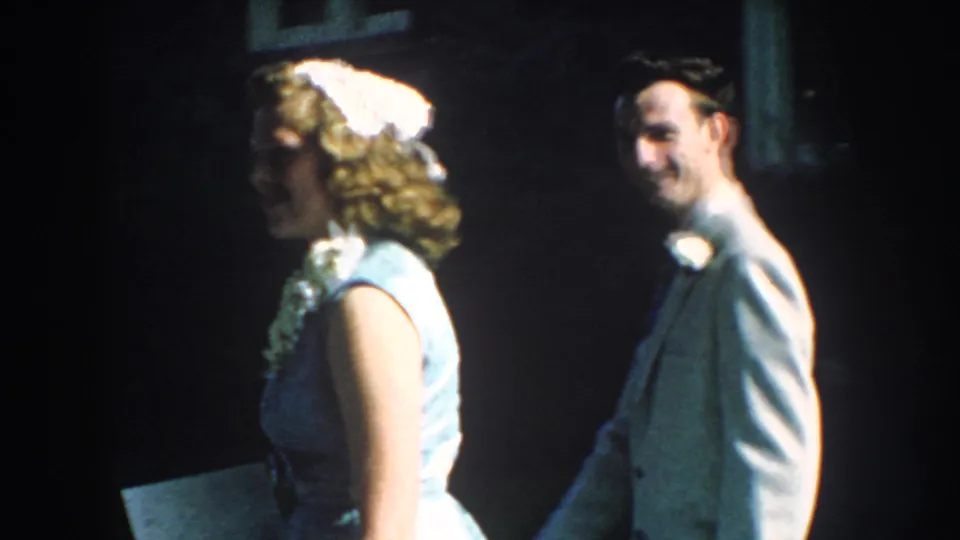 1959 Nebraska home-movie frame of a woman in a light blue dress and white headband near a smiling man in formal attire.