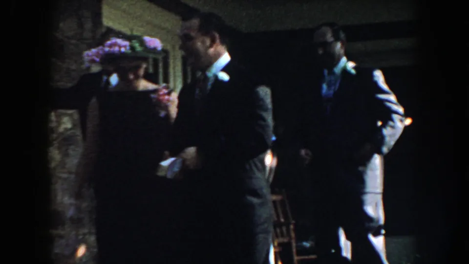 1959 Nebraska home-movie frame of formally dressed adult guests near a stone wall, with a floral hat and dark suits visible.