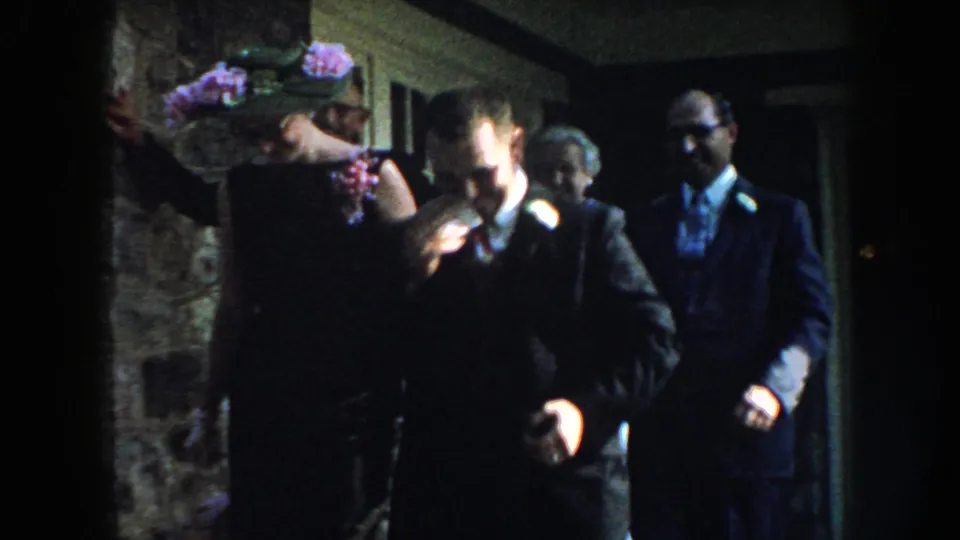 1959 Nebraska home-movie frame with a suited man in front and other formally dressed adults behind him in a dim interior.