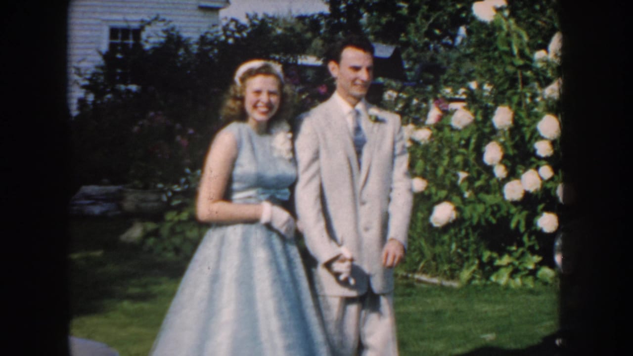 1959 Nebraska home-movie frame of a teenage girl in a light blue dress and a boy in a gray suit posing in a garden.