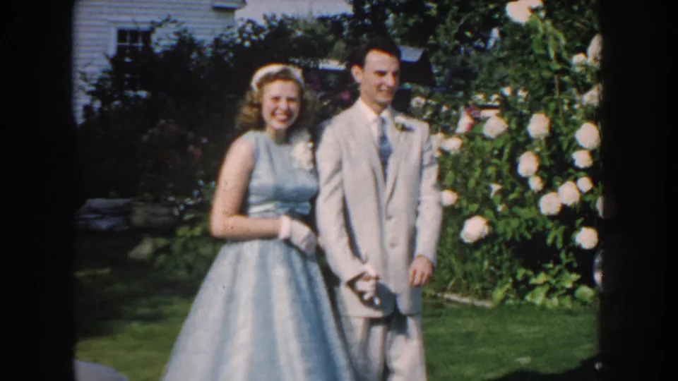 1959 Nebraska home-movie frame of a teenage girl in a light blue dress and a boy in a gray suit posing in a garden.