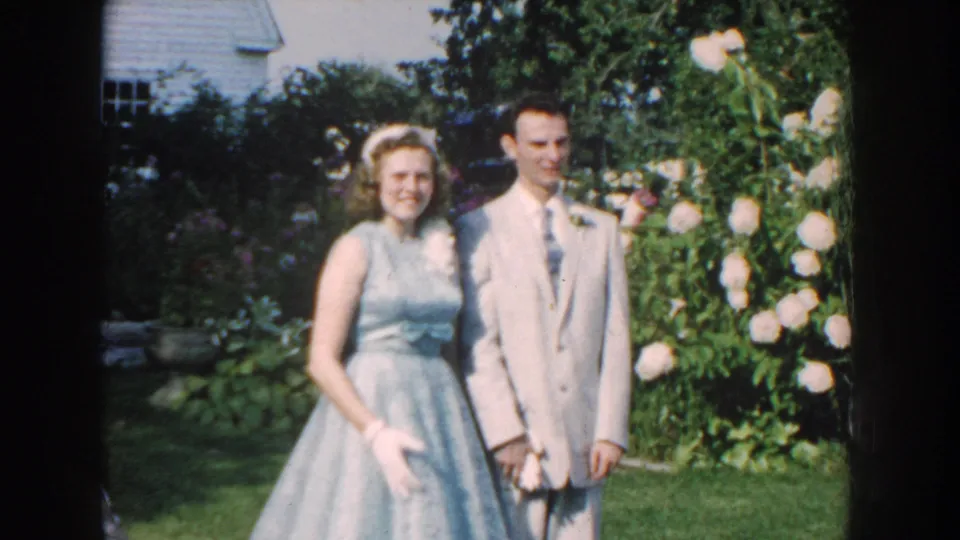 A 1959 Nebraska home-movie still shows a teenage couple in formalwear posing closely in a garden.