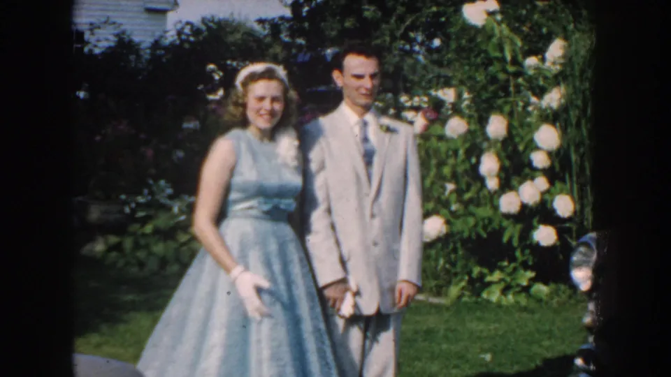 Teenage girl in a pale blue dress and boy in a gray suit pose outdoors in 1959 Nebraska home-movie footage.