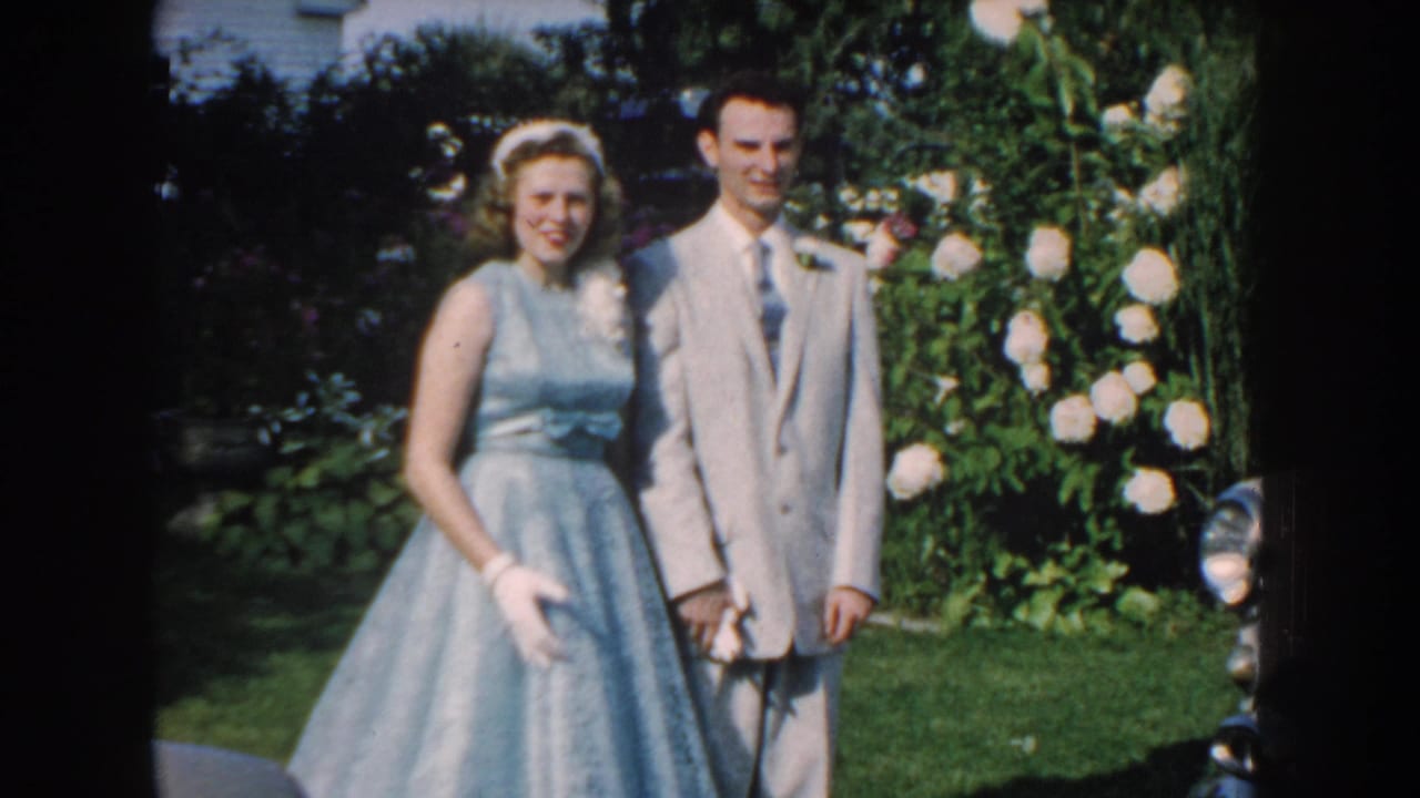 In 1959 Nebraska home-movie footage, a woman in a pale blue dress and a man in a light suit pose together in a sunlit garden.