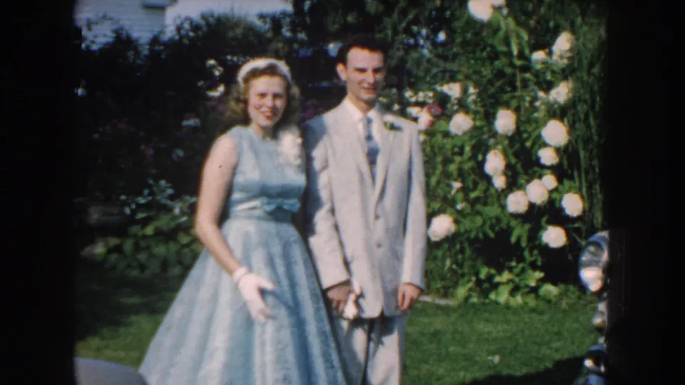 In 1959 Nebraska home-movie footage, a woman in a pale blue dress and a man in a light suit pose together in a sunlit garden.