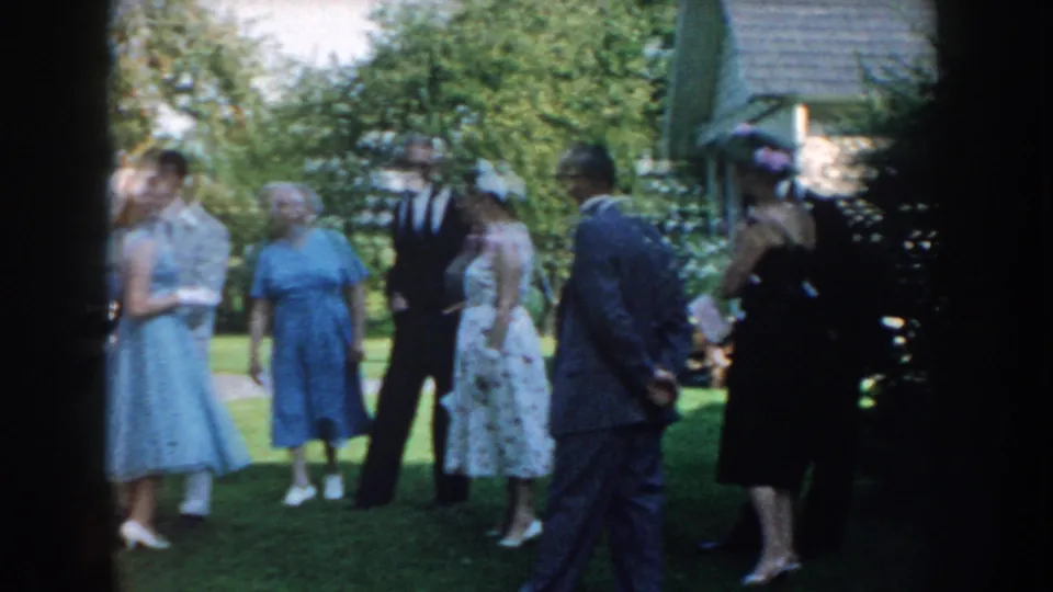Guests in mid-century dresses and suits gather on a Nebraska lawn, with the home-movie camera partially framed by dark film edges.