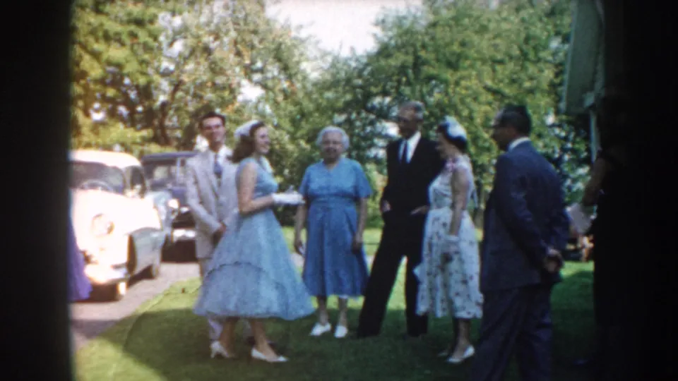 A small wedding group stands on a Nebraska lawn near parked mid-century cars, with the couple and older relatives visible among the guests.