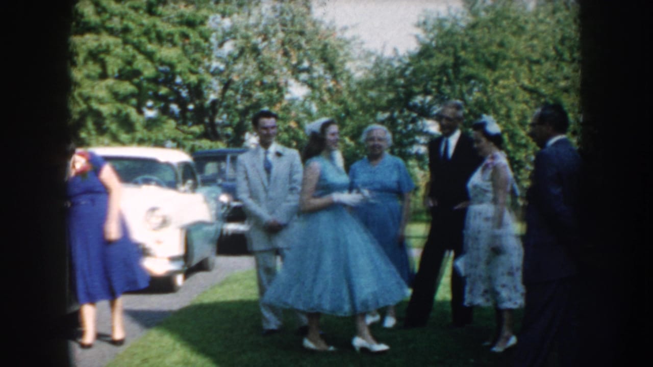 In Nebraska 1959 home-movie footage, formally dressed adults stand and talk on a lawn near several parked mid-century cars.