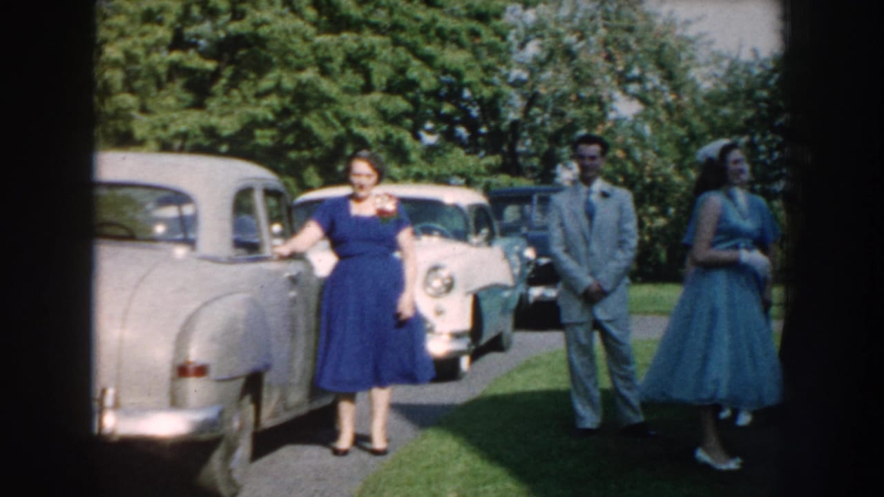 Woman in blue dress stands near a silver car with other guests nearby.
