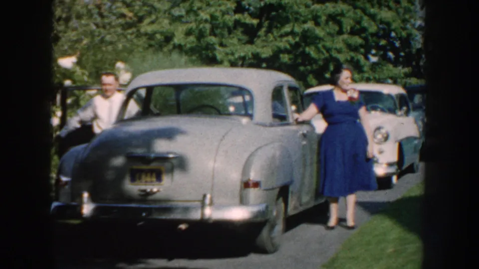 The rear of a silver mid-century car fills the foreground while a woman in blue and other adults stand near the driveway.