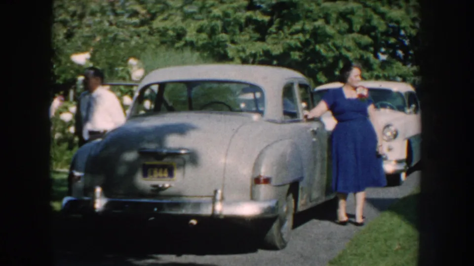 A formally dressed woman stands by a silver car on a grassy drive, with additional 1950s cars and trees behind her.