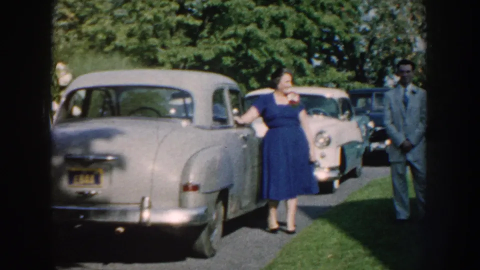1959 Nebraska home-movie frame showing a woman in a blue dress by a pale sedan, with suited adults and parked cars in a sunny garden.