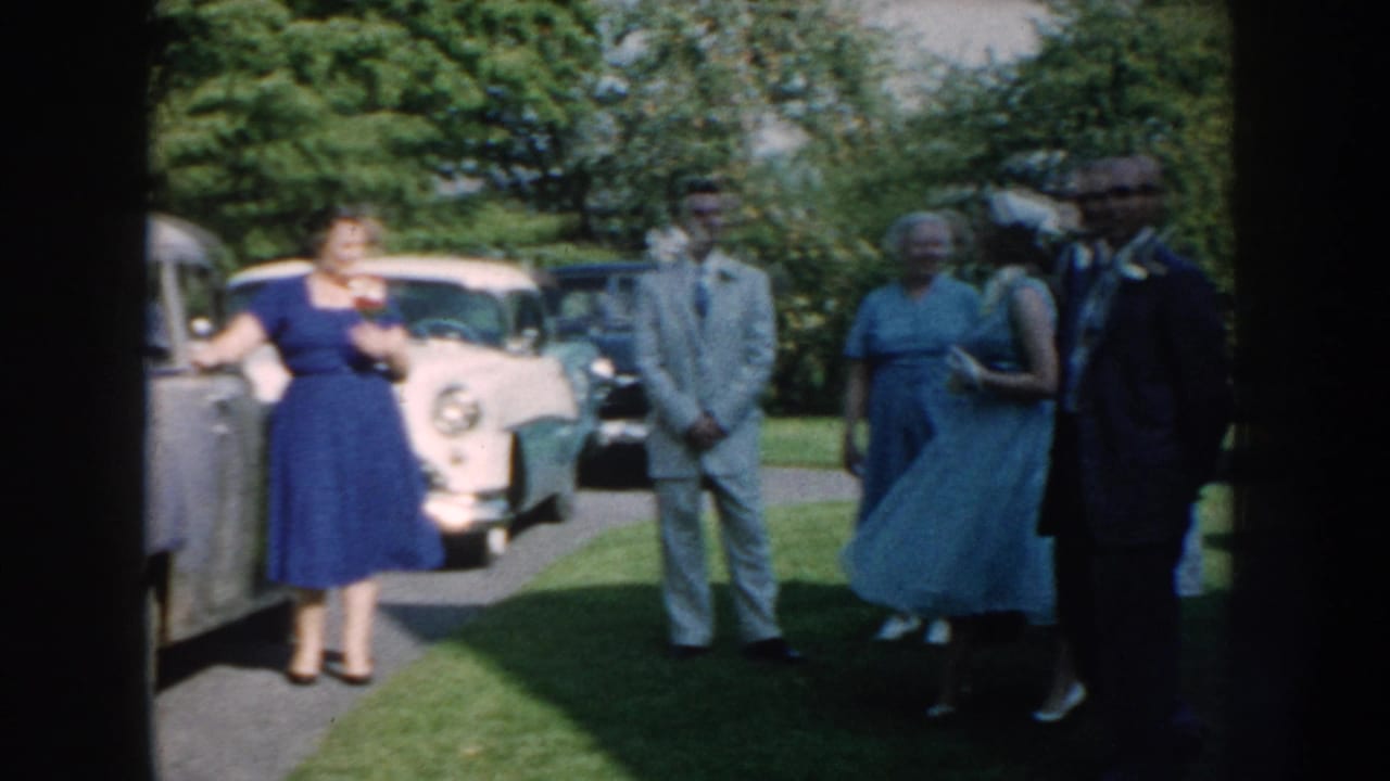 1959 Nebraska garden gathering with adults in dresses and suits standing near parked mid-century cars on a green lawn.