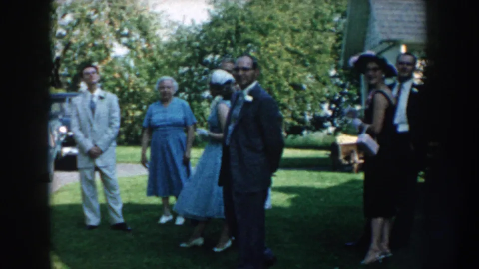 1959 Nebraska home-movie frame of adults in formal mid-century clothing gathered on a lawn, including hats, gloves, and handbags.