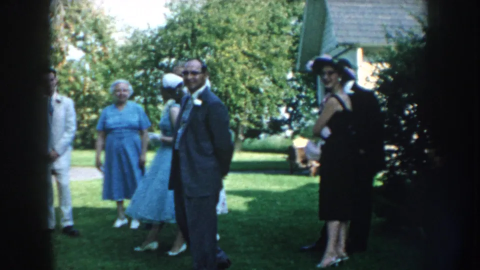 1959 Nebraska home-movie frame showing formally dressed adults on a lawn, with a man in a gray suit and a woman in a black dress and hat.