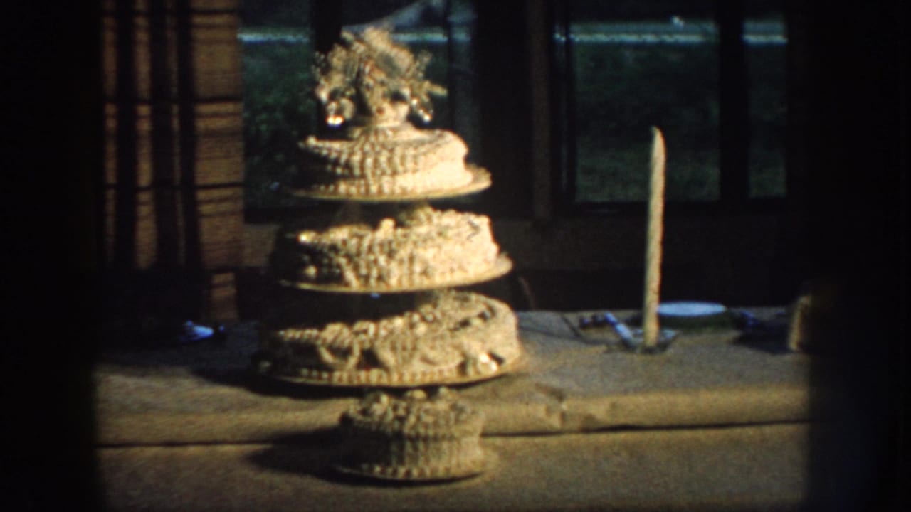 1959 Nebraska home-movie frame showing an ornate tiered birthday cake on a table near a tall candle and dark window.