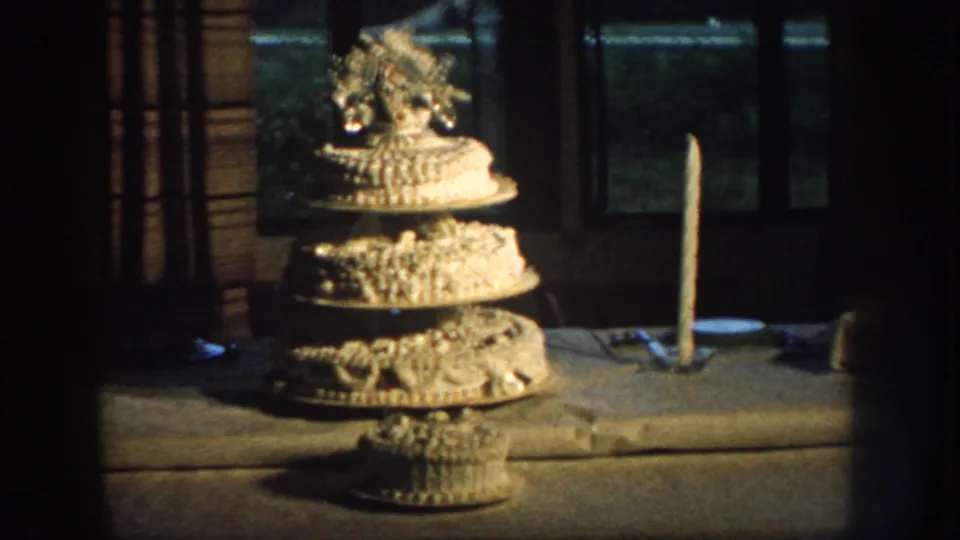1959 Nebraska home-movie frame showing an ornate tiered birthday cake on a table near a tall candle and dark window.