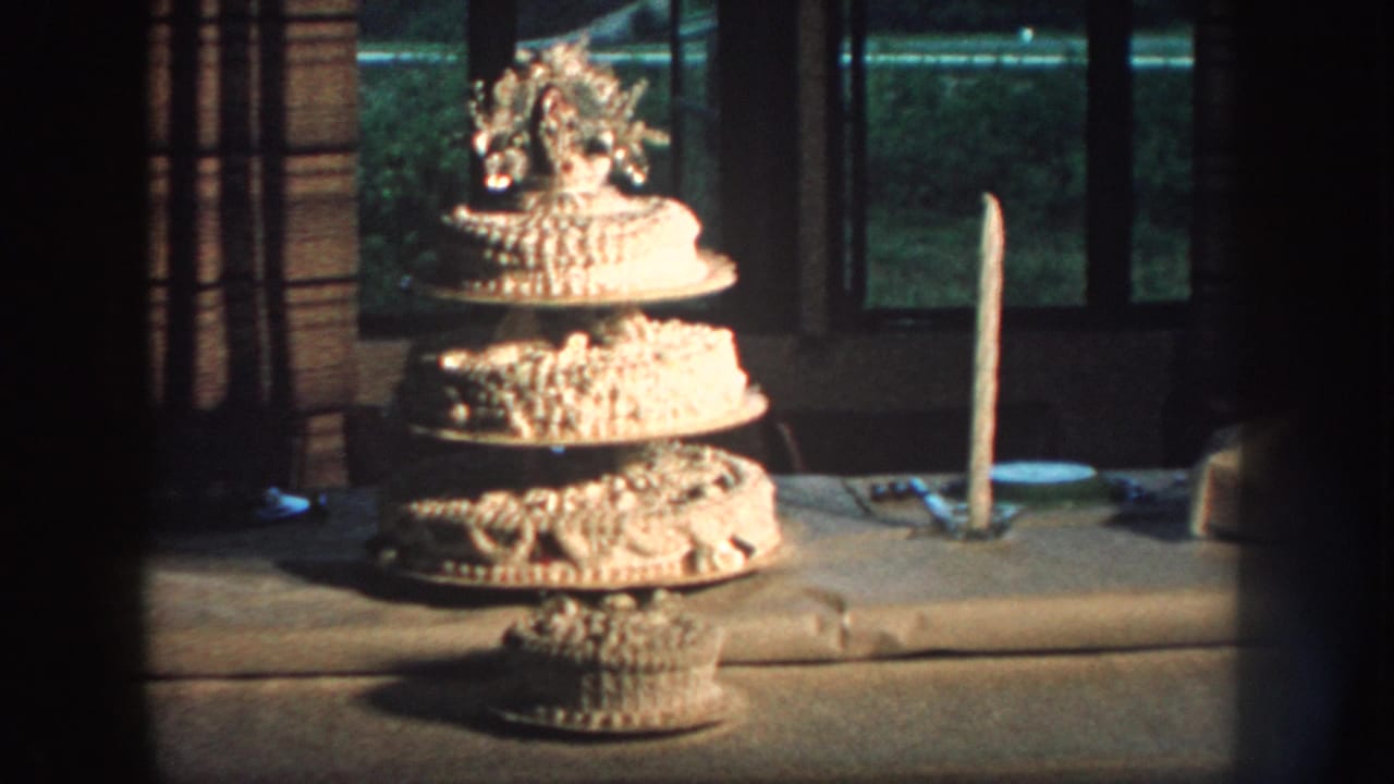 Home-movie still from Nebraska in 1959 with a multi-tier birthday cake, tall candle, and dim window light.