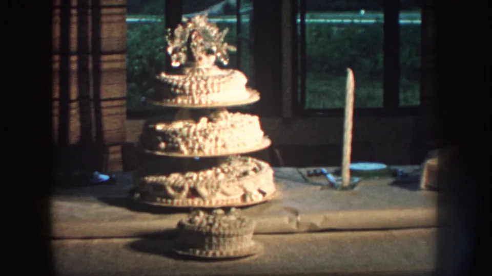 Home-movie still from Nebraska in 1959 with a multi-tier birthday cake, tall candle, and dim window light.