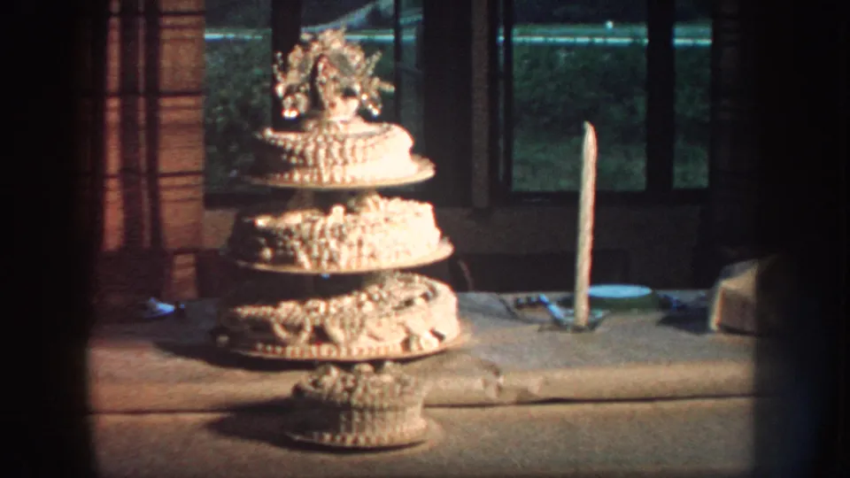 Soft 8mm frame of a Nebraska birthday cake in 1959, with a floral topper, tall candle, table, and window shadows.