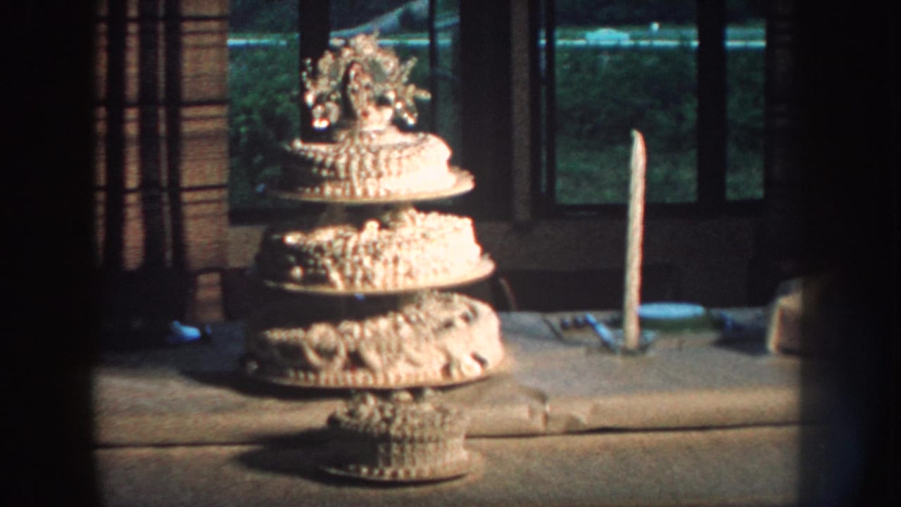 1959 Nebraska 8mm frame showing a decorated tiered white wedding cake on a stand with a tall candle and window behind it.