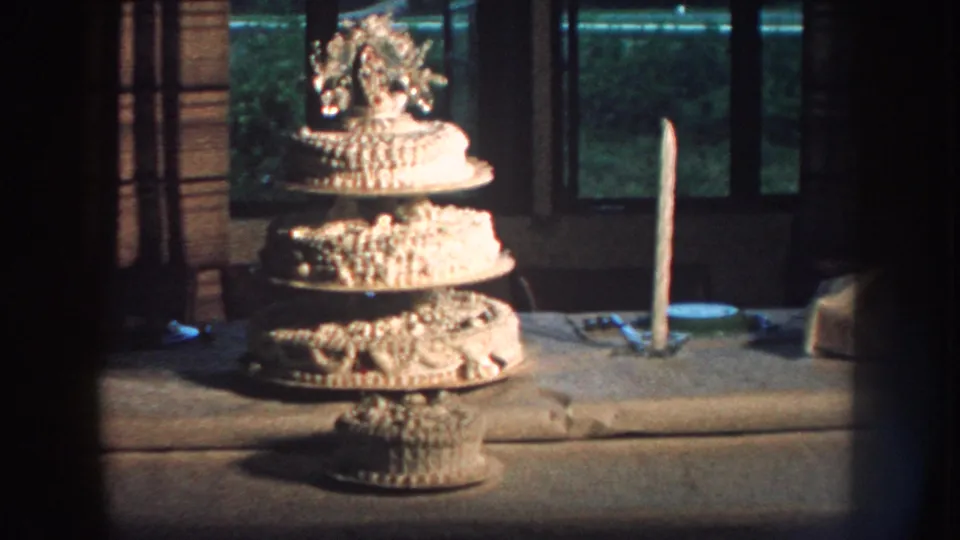 1959 Nebraska home-movie frame with the tiered wedding cake, tall candle, table setting, window, and greenery in view.