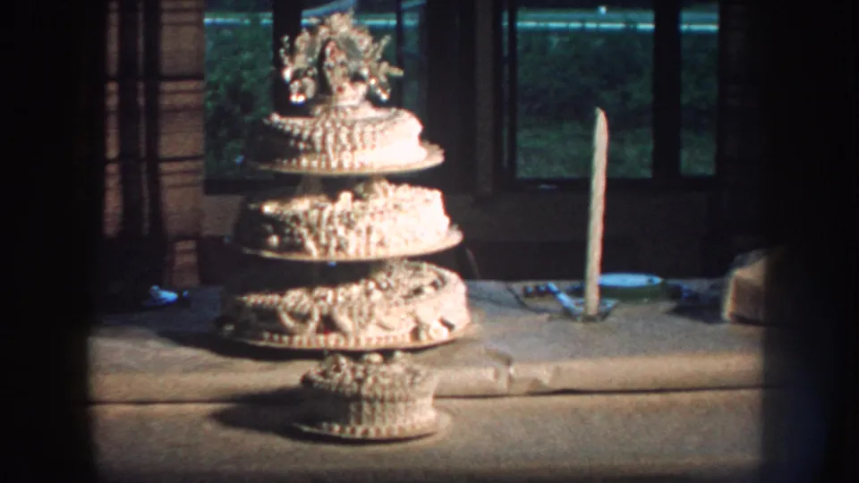 1959 Nebraska home-movie still of a white tiered wedding cake with floral topper, candle, tablecloth, and windowed background.