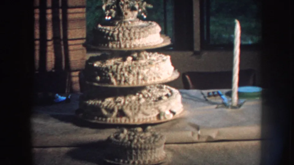 1959 Nebraska 8mm frame showing an ornate multi-tiered wedding cake on a table with a tall candle nearby.