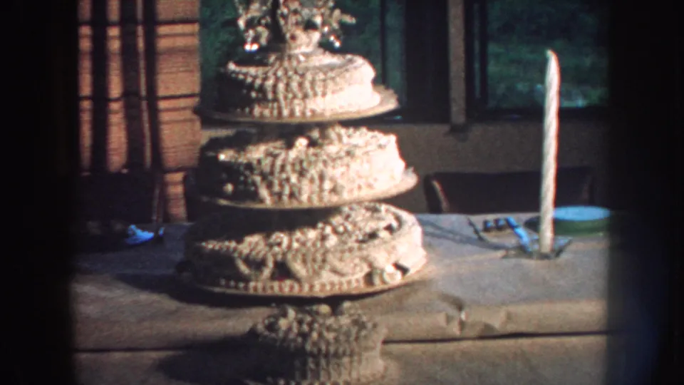 1959 Nebraska home-movie frame of a decorated tiered wedding cake with a candle and window in the background.