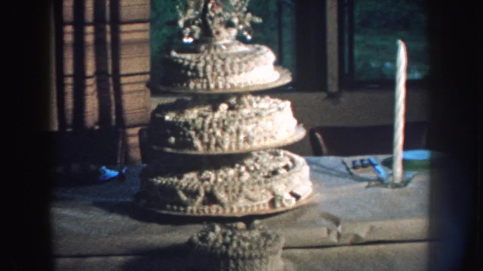 1959 Nebraska home-movie frame of a multi-tiered decorated wedding cake beside a tall candle in an interior setting.