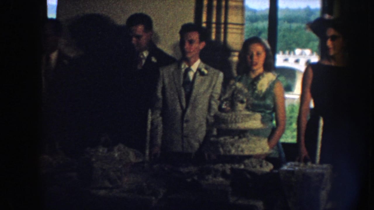 A bride and groom stand beside a tiered cake with guests nearby in a dim reception room.