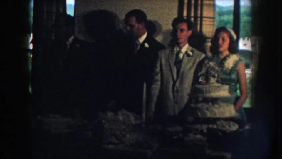 A 1959 Nebraska wedding reception frame shows the couple near the cake, partly obscured by shadowed guests in the foreground.