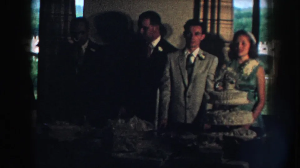 The final frame holds on the wedding couple and cake in a dim Nebraska interior, with guests visible in shadow nearby.