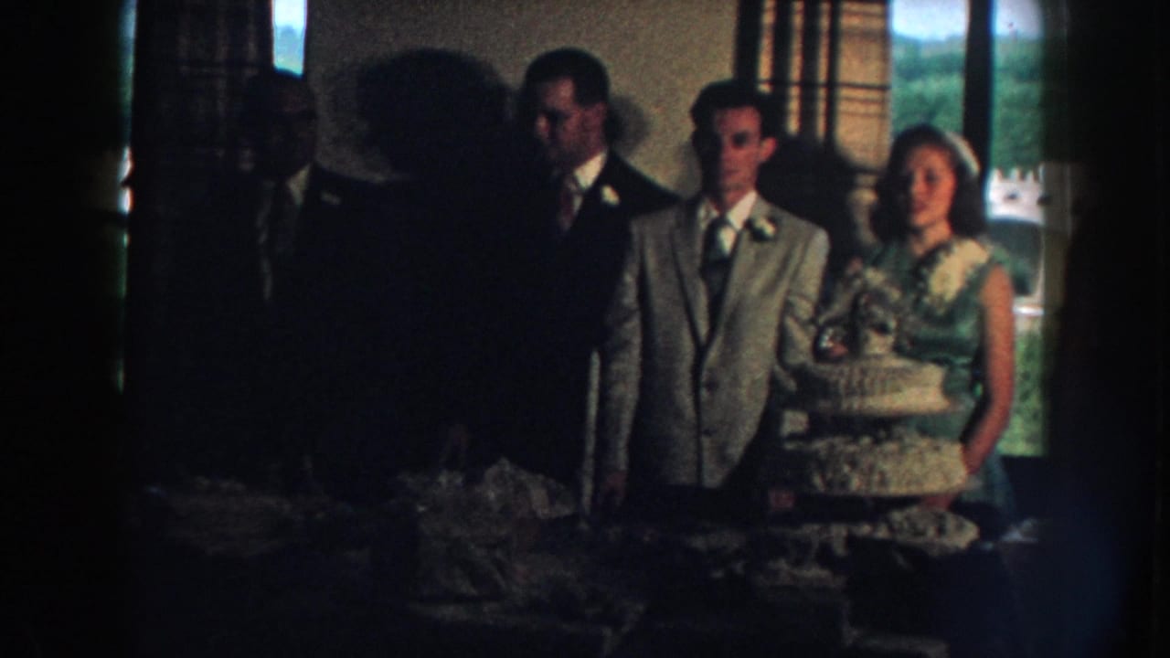 Wedding party stands near a tiered cake and reception table inside a room with large windows.