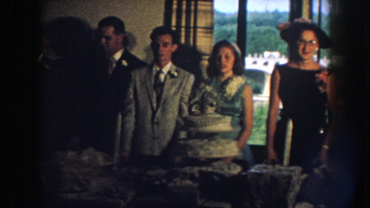 Bride and groom stand beside the cake while formally dressed guests line up near the window.
