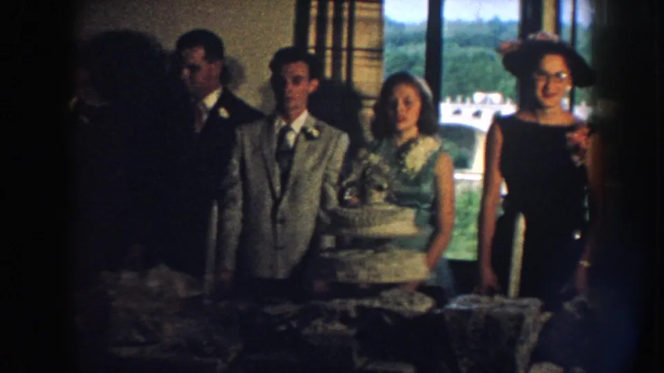 1959 Nebraska reception scene with a bride holding or standing beside a tiered cake, a groom, and guests in formal attire.
