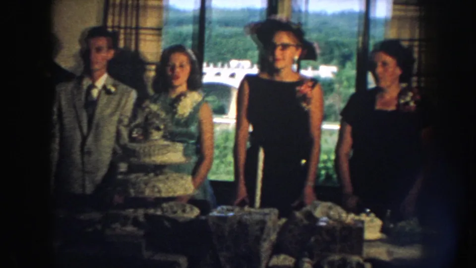 1959 Nebraska wedding reception frame showing the couple and guests in dresses and hats around a food-laden table.