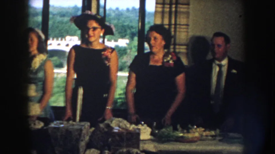 1959 Nebraska home-movie frame of wedding guests in formal wear standing behind a table of food near large windows.