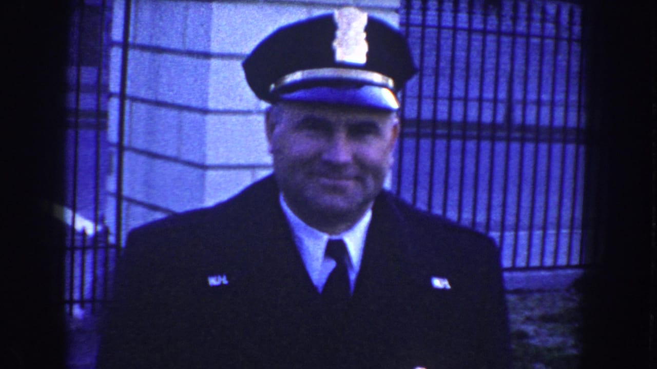 A smiling uniformed security officer faces the camera before a metal gate in 1960 Washington, D.C. home-movie footage.