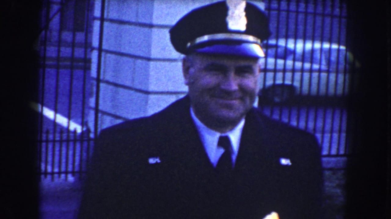 The uniformed officer smiles near a metal gate and stone building wall, captured in blue-shifted 8mm footage.