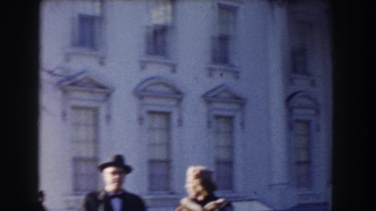 Man and woman in hats stand before a large white neoclassical facade.