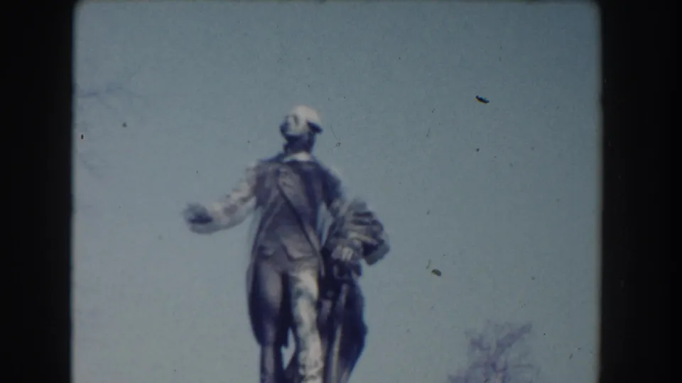 Visitor-shot 8mm frame of a large public statue in Washington, DC, seen from below with sky and faint tree branches behind it.