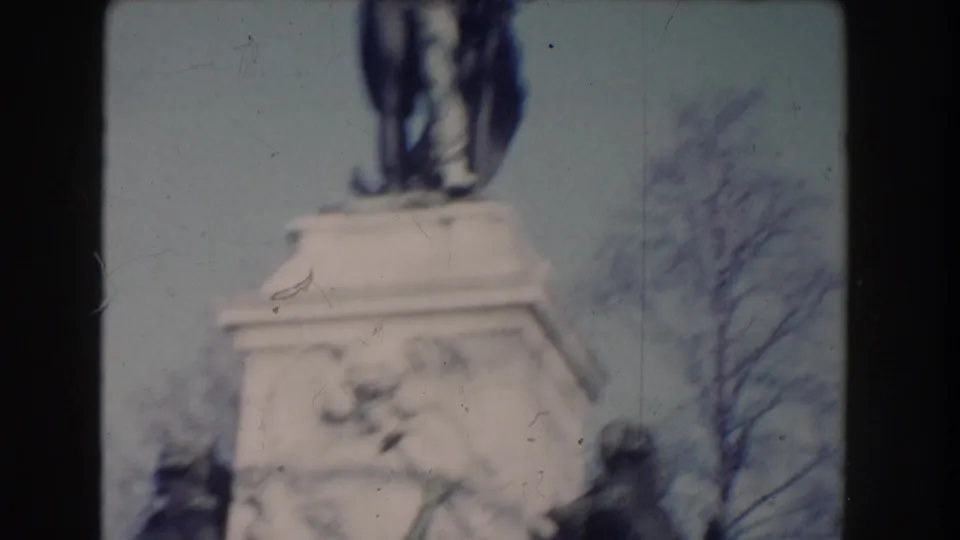 Soft 8mm view of a public statue pedestal in Washington, DC, with sculpted figures below and bare trees behind the monument.