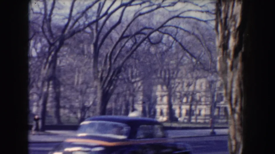 A 1960 Washington DC street view shows a dark American car moving past leafless trees with a large white building in the background.