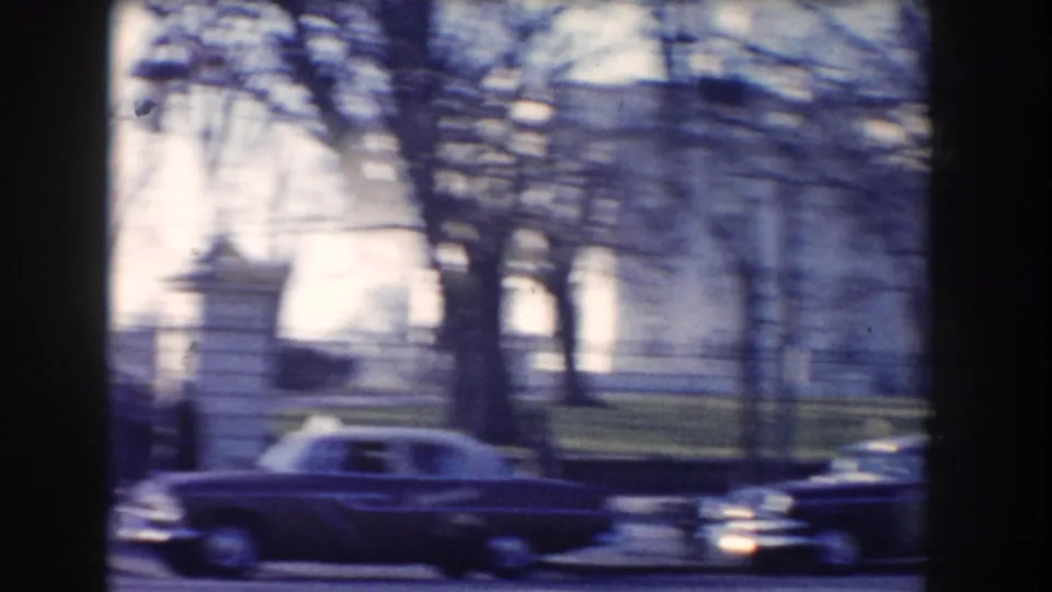 Two mid-century cars move along a Washington DC street near gates, bare trees, and a pale building seen through soft 8mm blur.